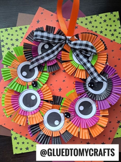 This cupcake liner Halloween wreath features orange, green, purple, and black liners with big googly eyes and mini black spiders. Finished with an orange ribbon and plaid bow, it pops against a festive Halloween-themed paper background.