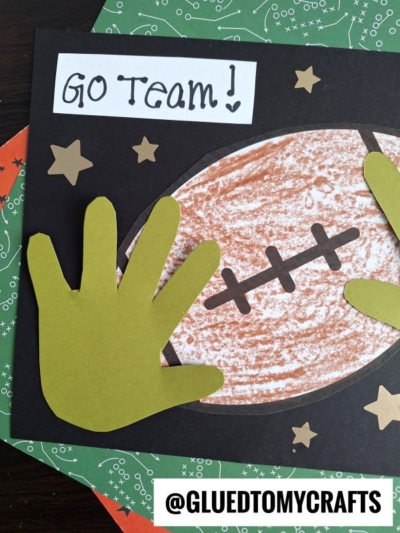 A handmade football keepsake card features a brown football, two green paper handprints, gold stars, Go Team! written on top, and the handle @gluedtomycrafts at the bottom, all set on patterned paper.