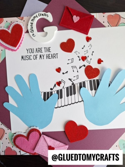 A handmade Valentines Day card featuring blue handprint piano keys on a keyboard, with red heart and music note cutouts. The message "You are the music of my heart" and @GLUEDTOMYCRAFTS appear at the bottom.
