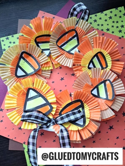 A charming candy corn wreath crafted from orange, yellow, and white cupcake liners with playful candy corn decorations, finished with a black and white plaid bow and set against colorful star-patterned paper in the background.