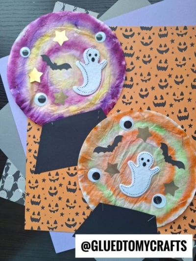 Two colorful coffee filter crystal ball globes with googly eyes, ghost and bat cutouts, and stars are displayed on orange pumpkin-patterned paper. The crafts have a Halloween theme.
