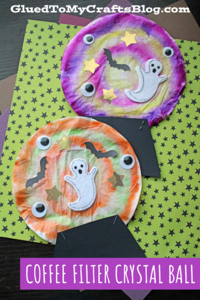Two colorful coffee filter crystal ball crafts, decorated with googly eyes, ghosts, bats, and yellow stars, sit on green star-patterned paper. Text reads: Coffee Filter Crystal Ball.