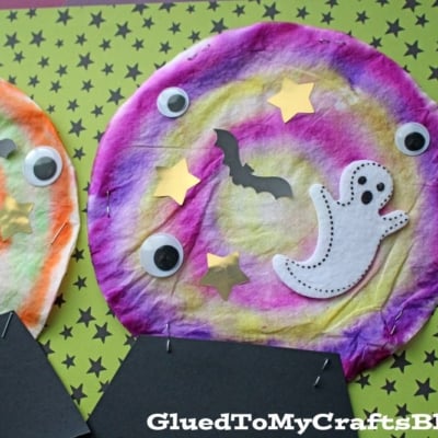Two colorful coffee filter crystal ball crafts, decorated with googly eyes, ghosts, bats, and yellow stars, sit on green star-patterned paper.