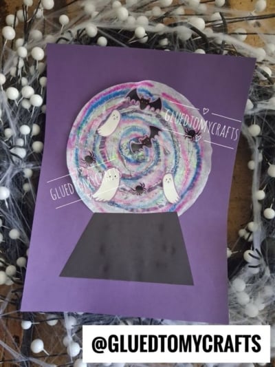 A coffee filter crystal ball craft on purple paper shows ghosts, bats, and spiders inside a colorful swirl. White berries and web-like material surround the scene. @GLUEDTOMYCRAFTS is written at the bottom.