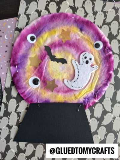 A Halloween craft featuring a paper snow globe with a ghost, bat, googly eyes, and gold stars—made to look like a magical coffee filter crystal ball—using a tie-dyed coffee filter and black paper, all set on a ghost-patterned background.