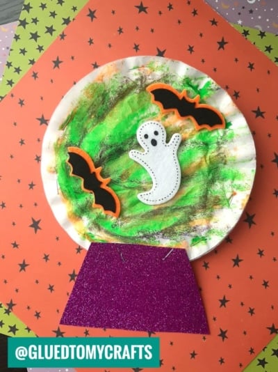 A paper plate craft features a ghost and two bats on a marbled green and orange background, set atop a purple glitter base, resembling a spooky coffee filter crystal ball snow globe. @GLUEDTOMYCRAFTS is in the bottom left corner.