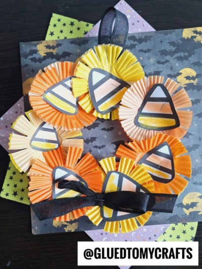 A festive candy corn wreath made from orange and yellow cupcake liners with paper candy corn cutouts, black ribbon, and decorative star paper in the background.