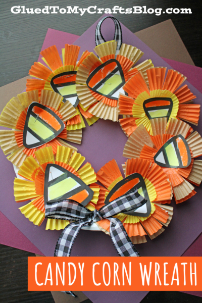 A festive candy corn wreath crafted from yellow, orange, and white cupcake liners and paper candy corn shapes, finished with a black-and-white plaid bow. The background showcases layered colored paper sheets.