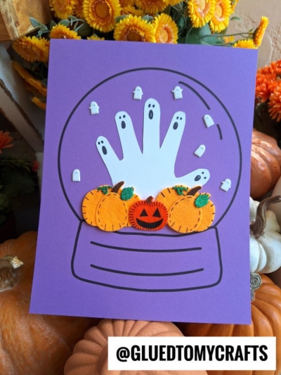 A purple card features a Halloween snow globe with a handprint ghost, surrounded by small ghost cutouts and felt pumpkins. The background includes orange flowers and real pumpkins. A label reads @GLUEDTOMYCRAFTS.