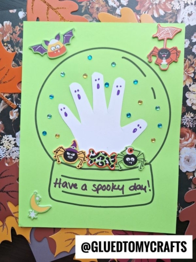 A green card shows a halloween snow globe made from paper, featuring a white handprint decorated as ghosts. Colorful stickers of spiders, a bat, a moon, and sequins surround it. Text reads, “Have a spooky day!” with a @GLUEDTOMYCRAFTS tag.