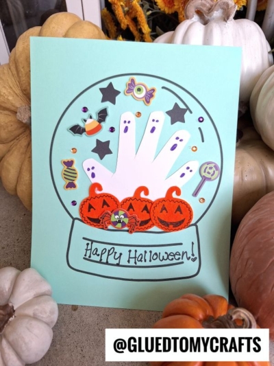 A Halloween snow globe craft featuring a handprint inside, decorated with smiling pumpkins, candy, sequins, and "Happy Halloween!" surrounded by pumpkins. @GLUEDTOMYCRAFTS is labeled at the bottom.