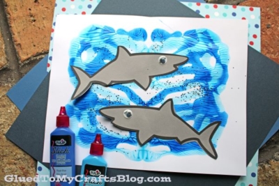 A handmade paint splat shark craft that features a gray paper shark with googly eyes on blue, on a splattered paint background