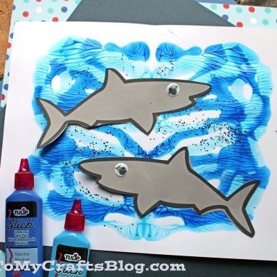 A handmade paint splat shark craft that features a gray paper shark with googly eyes on blue, on a splattered paint background