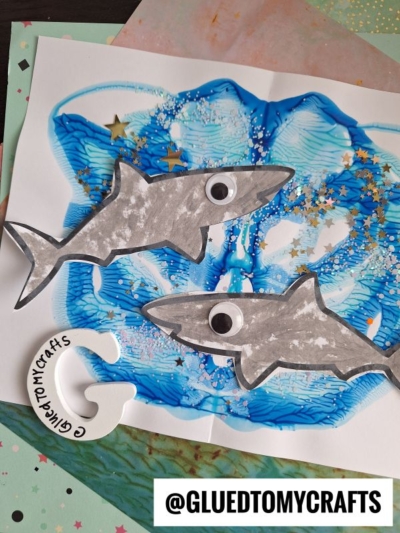 Two gray paint splat sharks with googly eyes rest on a blue and white painted background sprinkled with gold star confetti. A white letter G with Glued to My Crafts written on it sits nearby. @GLUEDTOMYCRAFTS is at the bottom.