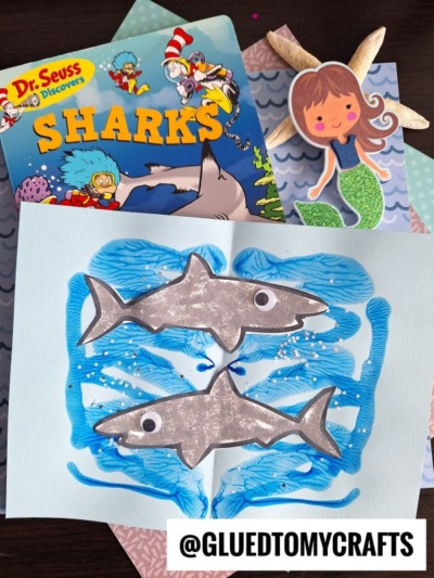 A children’s book titled “Dr. Seuss Discovers: Sharks,” a mermaid cutout, and an open paint splat shark craft with two painted sharks featuring googly eyes on blue waves and white speckles. The handle @GLUEDTOMYCRAFTS is at the bottom.