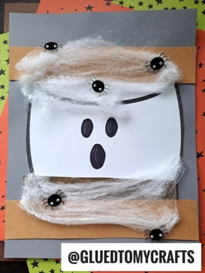 A Halloween craft featuring a ghost s'mores design made from white paper with black oval eyes and mouth, web-like cotton, and small black spiders on a gray and brown background. @GLUEDTOMYCRAFTS is written at the bottom.