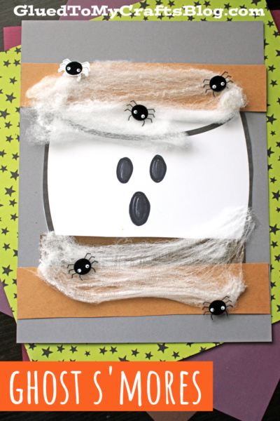 A Halloween craft card featuring a ghost face with black eyes and mouth, decorated with white cotton-like webs, ghost s'mores shapes, and small black spiders. The background is gray and brown with star-patterned paper underneath.