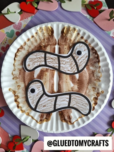 A paper plate with a paint splat folded to create a symmetrical pattern, featuring two playful paper worms with googly eyes, surrounded by heart-shaped cutouts on a striped background. @GLUEDTOMYCRAFTS is written at the bottom.