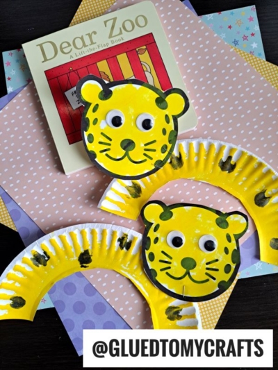 Two adorable paper plate leopards, painted yellow with googly eyes and black spots, are displayed on pastel patterned paper next to the children’s book Dear Zoo. A watermark reads “@GLUEDTOMYCRAFTS.”.
