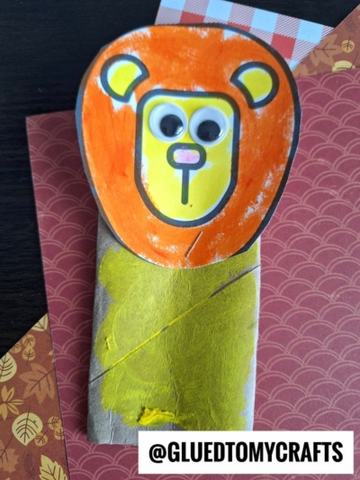 A toilet paper roll lion craft made from a painted roll, with a yellow face, orange mane, googly eyes, and a pink nose, sits on patterned paper. @GLUEDTOMYCRAFTS is written at the bottom.