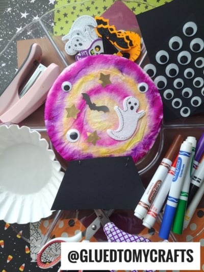 Colorful Halloween craft supplies and decorations, including markers, googly eyes, paper shapes, a stapler, a completed ghost and bat craft, and a whimsical coffee filter crystal ball with @GLUEDTOMYCRAFTS text at the bottom.