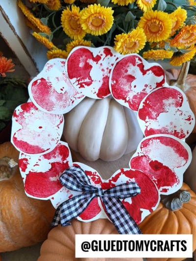 A fall wreath made of red and white painted circles, decorated with a black-and-white gingham bow, is displayed with white and orange pumpkins and yellow flowers. Reminiscent of an apple paper plate wreath, this festive creation brings a charming touch to your autumn décor. Text at the bottom reads @GLUEDTOMYCRAFTS.