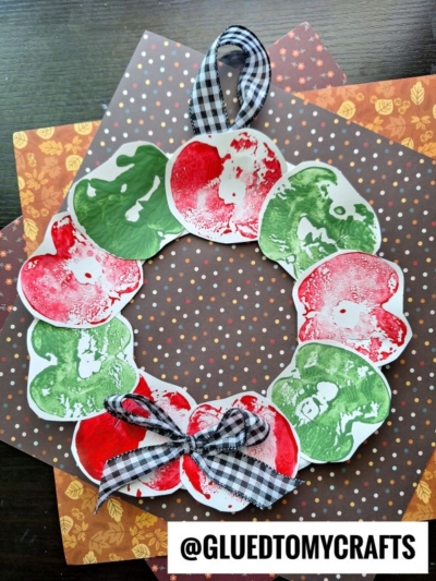 A festive apple stamped wreath made from red and green apple prints on paper, shaped into a circle. It’s topped with a black-and-white gingham ribbon bow and placed on autumn-patterned papers. The handle also features the same gingham ribbon.