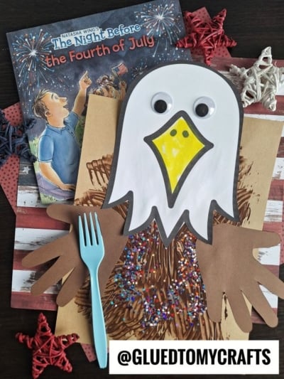 A children's bald eagle craft made with brown and white paper, googly eyes, and glitter sits on a book titled The Night Before the Fourth of July. Nearby are red star decorations and a blue fork perfect for fork painting crafts. @GLUEDTOMYCRAFTS is on the image.