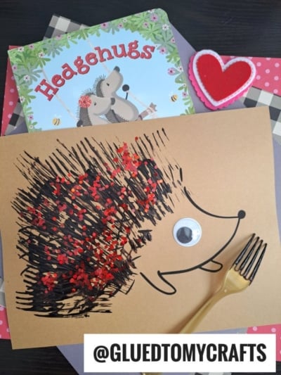 A handmade hedgehog craft using fork painting crafts with brown paper, black paint strokes, red glitter, a googly eye, and a plastic fork; beside a “Hedgehugs” book and heart-shaped decoration. @GLUEDTOMYCRAFTS is written at the bottom.