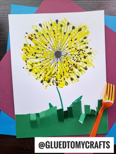 A vibrant dandelion art project made with yellow paint using fork painting crafts, featuring green paper grass and an orange plastic fork on colorful paper backdrops. The handle @GLUEDTOMYCRAFTS is shown at the bottom.