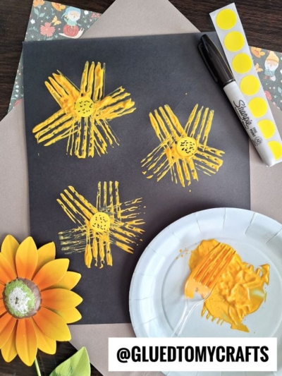A black paper displays four yellow paint flowers with textured petals, created using fork painting crafts. A Sharpie, yellow dot stickers, a painted paper plate, and a fake sunflower are nearby. The handle @GLUEDTOMYCRAFTS is at the bottom.