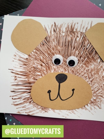 A handmade bear face craft made with fork painting crafts techniques for the fur, large paper ears, a paper snout with a drawn nose and mouth, and two googly eyes on white paper. @GLUEDTOMYCRAFTS is written at the bottom.