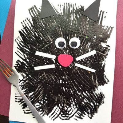 A black cat craft made with fork painting crafts—painted fork strokes on white paper, finished with googly eyes, paper whiskers, nose, and ears. A fork sits beside the artwork. Instagram handle @gluedtomycrafts is visible.
