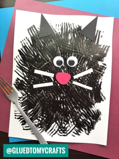 A black cat craft made with fork painting crafts—painted fork strokes on white paper, finished with googly eyes, paper whiskers, nose, and ears. A fork sits beside the artwork. Instagram handle @gluedtomycrafts is visible.
