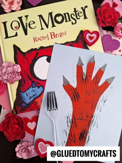 A Love Monster book, pink and red paper flowers, a fork painted monster hand art with gray claws on light blue paper. Heart decorations and the @GLUEDTOMYCRAFTS handle are also visible.