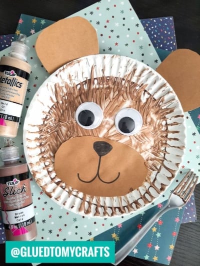 A bear face craft made from a paper plate painted brown, using fork painting crafts for texture, with added cardboard ears and snout, large googly eyes, and a black nose, surrounded by paint bottles and craft paper.