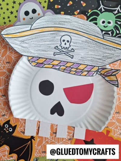 A paper plate pirate skull craft, inspired by a classic paper plate skeleton, features a striped hat, red eye patch, and skull-and-crossbones, surrounded by Halloween decorations like a bat, a spider, and spider webs.