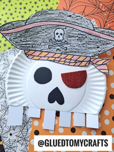 Pirate face craft made from a paper plate, with a colored pirate hat, orange eye patch, and skull details, giving it the spooky look of a paper plate skeleton, placed on Halloween-themed paper.
