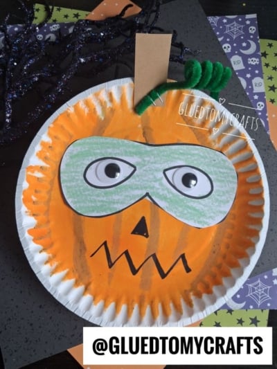 A Halloween craft of a superhero pumpkin face with a green mask and googly eyes, made from a painted paper plate, green pipe cleaners, and a brown paper stem. Halloween-themed paper decor is in the background. Text: @GLUETOMYCRAFTS.