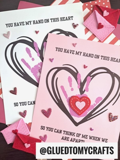 Two handmade Valentine’s cards feature a large heart outline with a child’s handprint keepsake inside and the words You have my hand on this heart so you can think of me when we are apart! Felt and glitter hearts are scattered around the scene.