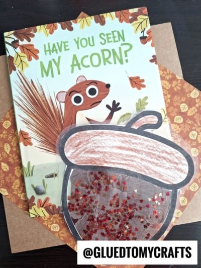 A children’s book titled Have You Seen My Acorn? featuring an illustrated squirrel sits on a desk. In front is a handmade acorn suncatcher craft decorated with brown coloring and red glitter. The handle @GLUEDTOMYCRAFTS is at the bottom.