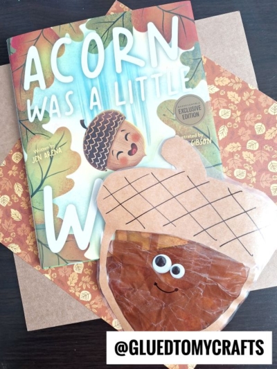 A children’s book titled Acorn Was a Little Wild sits next to an adorable acorn suncatcher craft with googly eyes and a smiling face, on a brown surface. @GLUEDTOMYCRAFTS is labeled at the bottom.