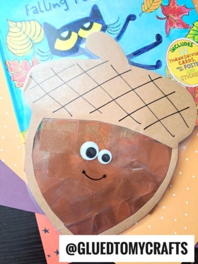A handmade acorn suncatcher craft with googly eyes and a smile, made from brown tissue paper and paper cutouts, sits on top of a children’s book featuring a black cat and autumn leaves. @GLUEDTOMYCRAFTS is labeled at the bottom.