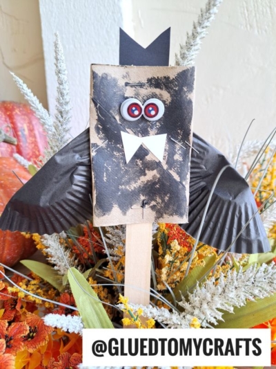 A toilet paper roll bat craft made from a paper bag, googly eyes, and black cupcake liners for wings, displayed among fall flowers. Text at the bottom reads @GLUEDTOMYCRAFTS.