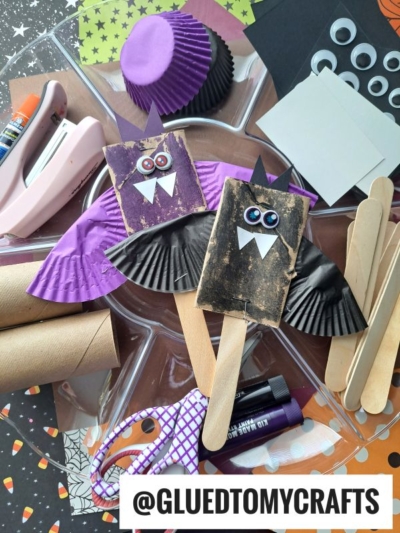 A craft project display features two bat puppets made from popsicle sticks, cupcake liners, googly eyes, and paper—resembling a toilet paper roll bat—surrounded by craft supplies like scissors, glue, cardboard tubes, and more. The handle @GLUEDTOMYCRAFTS is shown.