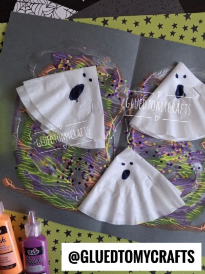 Three ghost crafts made from folded white paper and coffee filter ghost designs are placed on a colorful painted background, surrounded by craft supplies and star-patterned paper. @GLUEDTOMYCRAFTS is written at the bottom.