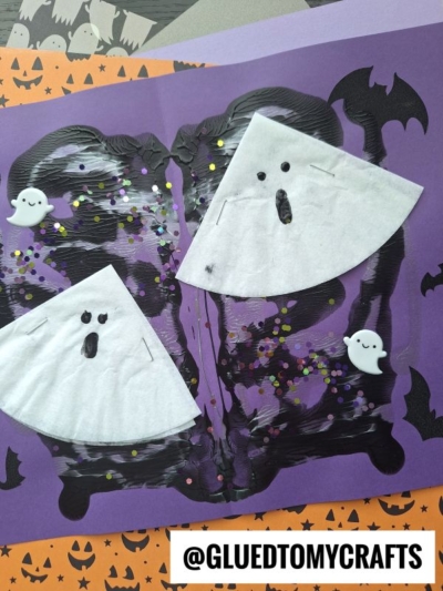 Two paper ghosts with shocked faces and foam ghost stickers are on purple and black Halloween-themed paper, decorated with bats and glitter. A cheerful coffee filter ghost adds extra charm to the spooky scene. The handle @GLUEDTOMYCRAFTS is at the bottom right.