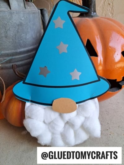 A charming cotton ball wizard craft with a blue paper plate wizard hat, fluffy cotton ball beard, and oval nose sits next to orange pumpkins. A white label reads @GLUEDTOMYCRAFTS.