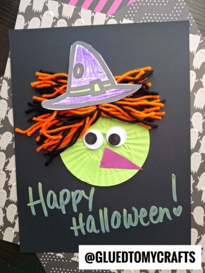 A Halloween witch craft with googly eyes, orange and black yarn hair, a purple hat, and a cupcake liner green witch face on black paper that says Happy Halloween! @GluedToMyCrafts is at the bottom.