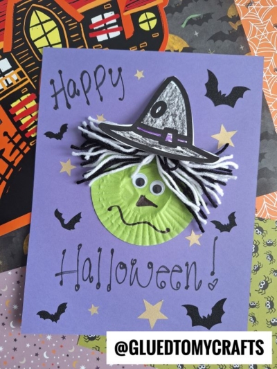A handmade Halloween card features a cupcake liner green witch with yarn hair, googly eyes, and a paper hat on purple paper adorned with bats, stars, and “Happy Halloween!” written across it. The handle @GLUEDTOMYCRAFTS is at the bottom.